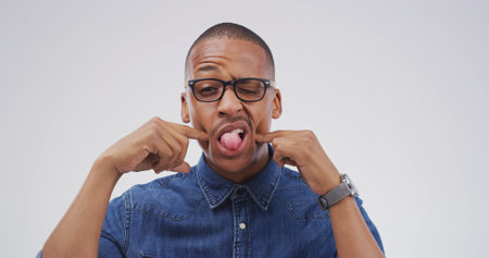 Portrait, funny face and black man in studio with silly facial expression on white background. Tongue out, mouth and African person with crazy gesture, emoji and playful comedy with glasses for comicの写真素材
