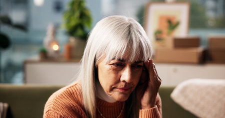 Mature woman, headache and home with stress on sofa for fatigue, strain or overthinking. Female person, vertigo or pain with migraine, discomfort or dizziness for mental health or insomnia in houseの写真素材