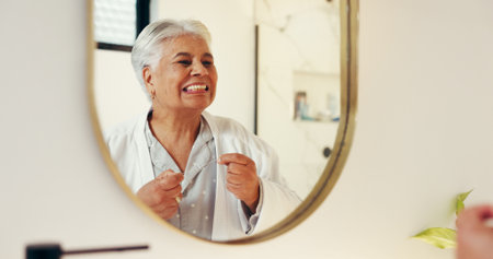 Dental health, mirror and senior woman with flossing in bathroom for morning routine in home. Oral care, reflection and elderly female person with string for cleaning teeth with hygiene in apartment.の写真素材
