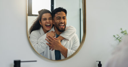 Couple, hug and laugh by mirror in bathroom for skincare, hygiene and happy together. Home, man and woman with reflection for beauty, funny joke and embrace for love with morning routine for groomingの写真素材