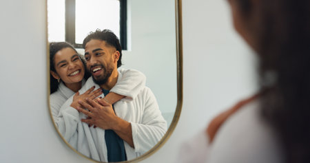 Couple, hug and mirror in bathroom for skincare, hygiene and laughing together. Home, man and woman with reflection for beauty, happiness and embrace for love with morning routine for groomingの写真素材