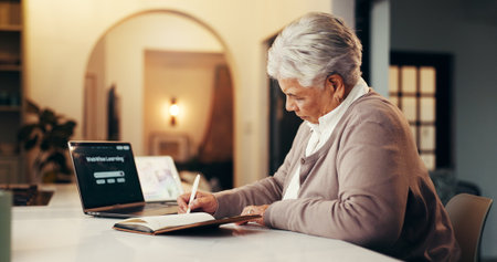 Senior woman, writing and elearning with laptop for online education, research or course in home. Elderly, female person or taking notes with book or computer at night for web or distance learningの写真素材