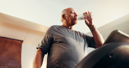 Elderly man, fitness and water with treadmill in home for workout, exercise or hydration. Active, below or senior person drinking mineral liquid on running machine for cardio break, rest or recoveryの写真素材