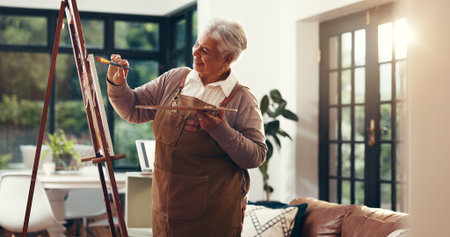 Senior woman, painting and happy in home with artwork, tools and canvas for inspiration in retirement. Elderly person, artist and smile with hobby, stroke and color with creative talent at houseの写真素材