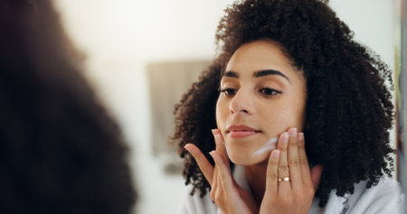 Beauty, lotion and mirror with woman in bathroom of home to apply facial product to skin. Cream, moisturizer and reflection with African person in apartment for cosmetics, dermatology or skincareの写真素材