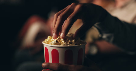 Hands, popcorn and snack at cinema in crowd for film screening, fast food and event for entertainment. Person, eating and relax for show, movies or container with audience at night in theaterの写真素材