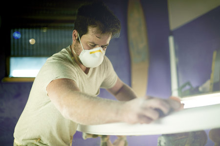 Craft, surfboard and polish with man in workshop for manufacturing, sports equipment and craftsman. Entrepreneur, maintenance and repair with person and mask for small business and quality assuranceの写真素材