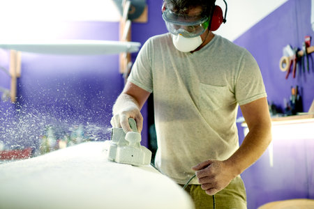 Workshop, man and tools for shape of surfboard, grinder and entrepreneur with skills for design or craft. Small business, artisan and person with machine for creation, manufacturing and polishingの写真素材