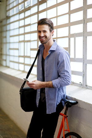 Man, portrait and phone in office with bicycle, networking and happy for eco friendly startup. Small business, entrepreneur and mobile at bike for campaign research, communication or green initiativeの写真素材