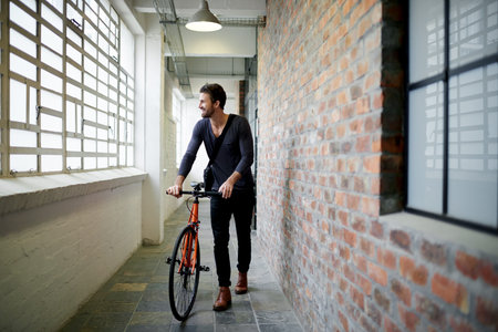 Man, bicycle and smile in hallway on travel by window, thinking or ready for ride at office building. Person, happy and bike in corridor for sustainability with eco friendly transportation on commuteの写真素材