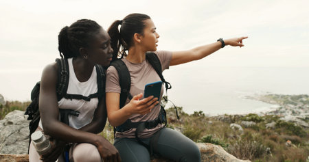 Girl, friends and hiking with phone on mountain, talk or point with digital map on trail. Women, discussion and smartphone for route, trekking and app for adventure, location or landmark in summerの写真素材