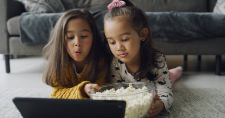 Children, sister or watching with tablet in home for online entertainment or subscription. People, girl or siblings streaming with technology or popcorn for movie, snack or bonding together on floorの写真素材
