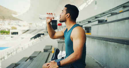 Stadium, man and drink water with earphones for fitness, hydration and running break. Outdoor, athlete person and relax with liquid beverage for training podcast, exercise and music for workout restの写真素材