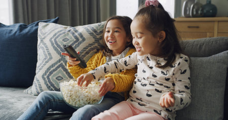 Popcorn, smile and watching TV with girls on sofa in living room of home together for streaming. Eating, relax and remote with sister children in apartment for bonding or television entertainmentの写真素材