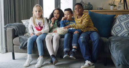 Happy kids, friends and watching tv with popcorn on sofa for movie or online entertainment. People, group or children eating snack with show for streaming subscription, cartoon or series in homeの写真素材
