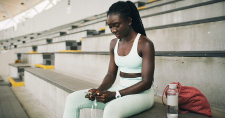 Stadium, black woman and typing with smartphone for fitness, online communication and scroll on app. Outdoor, female person and relax with mobile for chat message, social media and break for exerciseの写真素材