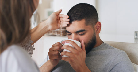 Sick, man and fever with drink at house for temperature, virus problem and flu symptoms. Couple, check forehead and monitor illness of male person, viral infection and herbal tea for recovery remedyの写真素材