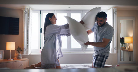 Couple, happy and pillow fight on bed with love, playful and relationship development in home. Man, woman and cushion war in bedroom for silly game, bonding and excited with energy in apartmentの写真素材