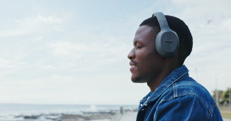 Travel, man and relax with headphones at ocean for peaceful trip, vacation playlist or mindfulness. Sky space, black person or listening to music at beach with thinking, holiday reflection and audioの写真素材