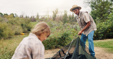 Man, woman and pitching tent in nature for camping, vacation adventure and bonding together. Couple, partner helping or setup equipment as outdoor shelter, campsite preparation and wilderness holidayの写真素材
