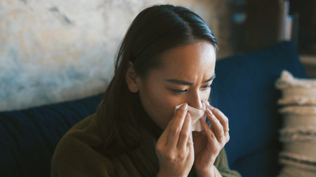Cold, woman and sick with sneeze on sofa from viral infection, disease and flu recovery. Respiratory illness, person and blowing nose in tissue with nasal congestion or healing from influenza in homeの写真素材