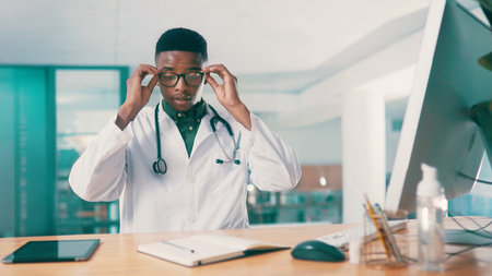 Computer, glasses and notebook with doctor black man at desk in hospital for results review. Healthcare, medical and research with medicine professional in office at clinic for agenda or scheduleの写真素材
