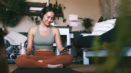 Music, phone and yoga with woman in living room of home for holistic or wellness routine. App, communication and headphones with Asian person on apartment floor for social media post or updateの写真素材