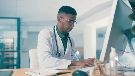 Black man, doctor and typing on computer for wellness, telehealth report or insurance records. Desktop, serious and medical worker with healthcare history, clinic schedule and research in officeの写真素材