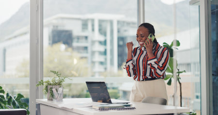 Phone call, fist pump and black woman with laptop for celebration, success and good news. African person, consultation and excited in workplace with promotion, achievement and customer service goalsの写真素材