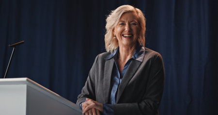 Podium, politics and portrait of mature woman on stage for democracy, election or government campaign. Microphone, smile and speech with speaker at conference, meeting or summit for support or voteの写真素材