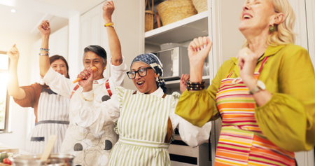Mature, happy women and dancing with food in kitchen for social gathering or celebration in home. Group, female people or friends with smile, vibe or vegetables for cooking success together in houseの写真素材