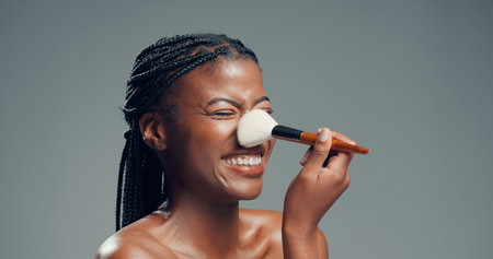 Makeup, beauty and happy black woman with brush in studio for shine, foundation or glow. Smile, apply cosmetics and model with product for facial makeover, change and powder on gray background spaceの写真素材