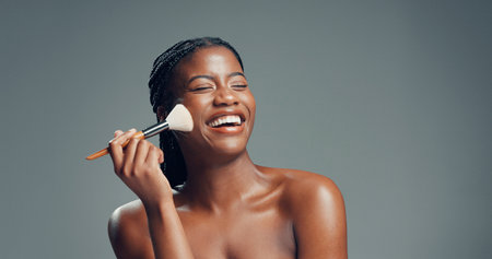 Makeup, beauty and black woman laugh with brush in studio for shine, foundation or glow. Funny, apply cosmetics and model with product for facial makeover, change and powder on gray background spaceの写真素材