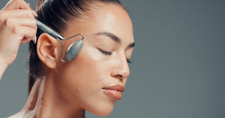 Woman, jade roller and massage in studio with skincare for beauty, dermatology and facial tension. Relax, person and cosmetic tool for lymphatic drainage, wellness or space on gray background.の写真素材