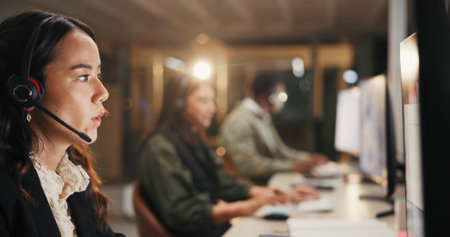 Woman, call center and helpdesk with headset at night for customer service or consulting at office. Female person, employee or agent talking for communication, typing and overtime at workplaceの写真素材