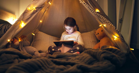 Night, child and reading in fort with tablet, fairy lights or ebook for fantasy bedtime story. Late, girl or relax with digital tech in home for online fairytale, kids storytelling app or literature.の写真素材