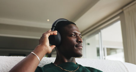 Home, headphones and black man on couch, thinking and listening to music, sound and podcast. Playlist album, relax and African person on sofa, audio and radio with contemplation and daydreamingの写真素材