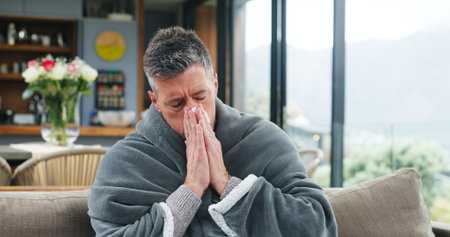 Blowing nose, sick and cold with man on sofa for virus infection, disease recovery and healthcare. Hayfever symptoms, influenza illness and healing with person and blanket in living room of homeの写真素材