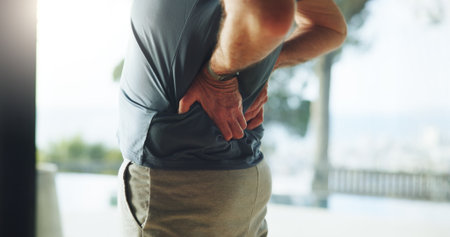Hands, senior man and back pain in fitness with muscle tension, workout accident and inflammation. Injury, old person or spine fracture in home from herniated disc, joint ache or training mistakeの写真素材