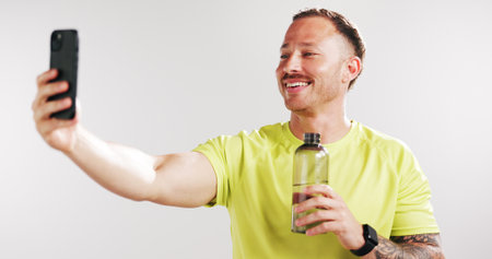 Selfie, water and fitness with man in studio for social media gym membership promotion and memory. Profile picture, personal trainer post and photography with person and bottle on white backgroundの写真素材