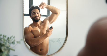 Man, spray armpit and bathroom mirror for hygiene in home, fragrance or reflection for morning routine. Person, grooming and shirtless with deodorant for odor, sweat and happy with bottle at houseの写真素材