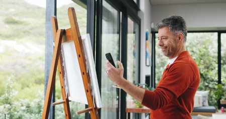 Painting, phone and picture with man in workshop for design reference, artist project and contact. Illustration inspiration, gallery artwork and canvas with mature person in studio for photographyの写真素材