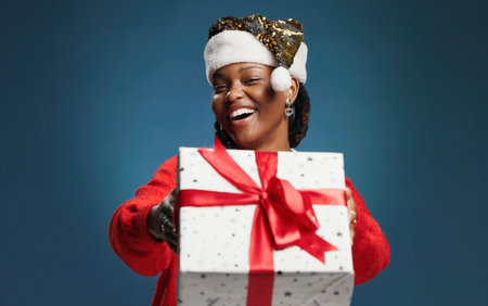 Christmas, gift box and portrait with African woman on studio space for celebration or surprise. Festive, giving and present with excited black person isolated on blue background mockup for traditionの写真素材