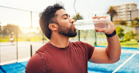 Man, drinking water and break at tennis court, sports and hydration for wellness with exercise routine. Person, athlete and bottle for electrolytes, training or fitness with rest for game in cityの写真素材