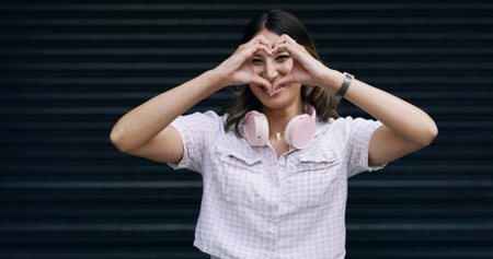 Happy woman, portrait and sign with heart hands in studio for love on a dark background. Female person, model or gen z with headphones, shape or emoji for romantic gesture, support or care on spaceの写真素材