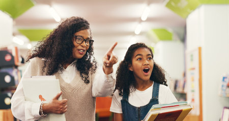 Teacher, kid and pointing in library with surprise for book collection, literature interest and study. Education, tutor or girl child with shock at school for literacy resources or academic knowledgeの写真素材