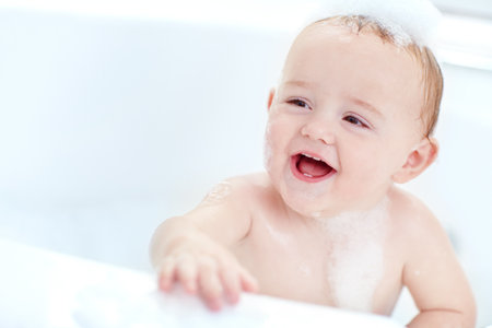 Bubbles, bathtub and baby laughing in bathroom for playing, washing and clean routine at home. Skincare, happy and cute boy toddler in water with foam for hygiene with growth and development in houseの写真素材