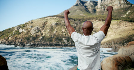 Runner, beach and celebration with man for fitness goal, success or win at sea. Back, sports achievement and mature athlete by ocean for workout target, health and earphones for music outdoorの写真素材