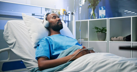 Black man, relax and sleep with headphones in hospital bed for streaming music, podcast and recovery. Male person, inpatient and listening to audio of playlist, album and healing nap from operationの写真素材