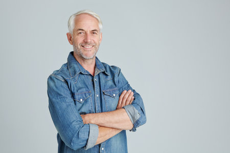 Portrait, man and smile in studio with arms crossed for casual fashion, positive attitude or wellness. Confident, happy or mature person on gray background with denim shirt, optimism or mockup space.の写真素材